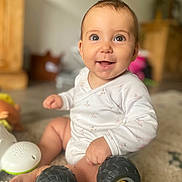 Elena a rejoint le concours — aidez-le/la à gagner de superbes lots ! baby, smiling, toy_car, carpet, indoors, child, hand, foot, face, clothing, white_outfit, happy, sitting, play, wheel, furniture, blurred_background, cute, infant, light
