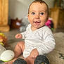 baby, smiling, toy_car, carpet, indoors, child, hand, foot, face, clothing, white_outfit, happy, sitting, play, wheel, furniture, blurred_background, cute, infant, light
