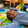 baby, child, play_mat, toy, giraffe_toy, colorful, nursery, infant, smiling, tongue_out, blue_clothing, quilted_jacket, floor, indoor, crawling_mat, cute, happy, young_child, person, closeup