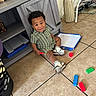 toddler, child, floor, tiles, blocks, toys, sneakers, green_clothing, cabinet, vase, dried_flowers, indoor, play, curious, messy, expression, person, hair, sitting, red_block