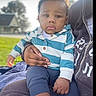 baby, child, person, outdoor, hoodie, striped_clothing, lap, hand, grass, house, trees, blurred_background, casual_clothing, cute, sitting, portrait, young_child, expression, blue_pants, sunlight