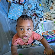 Antoine is registered to the contest to win money with this photo: toddler, child, playpen, blanket, pillow, stuffed_toy, toy, play_area, curious, indoors, looking_up, braided_hair, pink_shirt, baby, floor, carpet, learning_toy, colorful, cute, grip