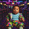 baby, child, arcade, retro, neon_lights, colorful, gaming, arcade_machines, vibrant_clothing, 90s_style, sneakers, floor_pattern, indoor, portrait, happy, smiling, sitting, playful, young_child, bright_colors