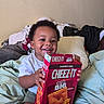 Antoine is registered to the contest to win money with this photo: toddler, child, smiling, cheezit_box, snack, bed, blanket, pillow, clothing, indoors, happy, person, curly_hair, sitting, holding, food, casual, home, young_child, comfortable