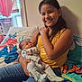 child, baby, smiling, girl, newborn, bedroom, window, curtains, air_conditioner, jeans, yellow_top, dinosaur_onesie, pillow, colorful_bedding, family, indoor, holding_baby, happy, caring, portrait