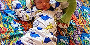 Matias is registered to the contest to win money with this photo: baby, sleeping, pajamas, dinosaur, plush_toy, blanket, colorful, pattern, floor, pacifier, infant, cute, cozy, child, toy, soft, clothing, resting, indoor, headwear