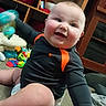 baby, child, infant, smiling, laughing, portrait, indoor, playroom, carpet, toys, sitting, black_clothing, orange_clothing, diaper, happy, face, eyes, hands, shelves, home