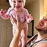 arm, baby, beanie, child, expression, family, happy, holding, home, indoor, light, love, man, onesie, person, playful, portrait, skin, smiling, tongue_out