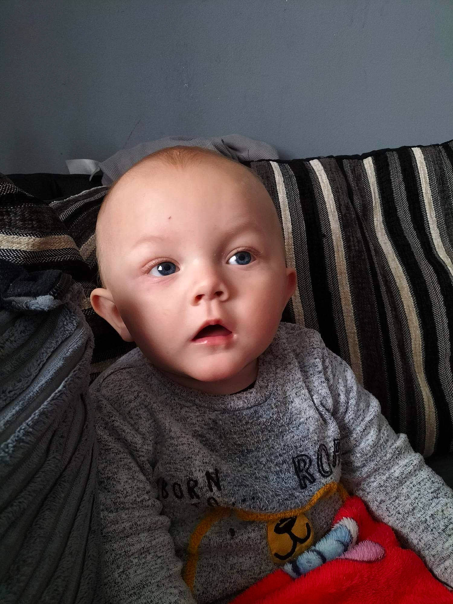 Sebastian is registered to the contest to win money with this photo: baby, baby_toddler_clothing, cheek, child, comfort, flash_photography, flooring, fun, grey, iris, linens, lip, nose, outerwear, pattern, person, sitting, skin, sleeve, toddler