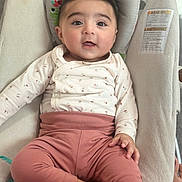 Fatima joined the competition — help win amazing prizes! baby, child, infant, sitting, clothing, pink_pants, white_top, face, cute, indoors, cushion, seat, hand, foot, expression, portrait, young, comfortable, looking, person