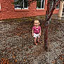 child, toddler, blonde_hair, tree, gravel, outdoor, building, window, pink_shirt, white_shorts, sandals, curious, standing, nature, sidewalk, leaves, daylight, person, young_child, casual_clothing