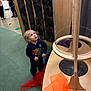 toddler, child, indoor, museum, interactive_exhibit, wooden_structure, scarf, curious, blonde_hair, green_floor, adult, conversation, yellow_wall, casual_clothing, educational, play_area, person, looking_up, standing, curiosity