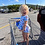 child, toddler, shopping_cart, barefoot, blonde_hair, parking_lot, sunny, outdoor, playful, tongue_out, clothing, shorts, top, vehicle, trees, blue_sky, clouds, metal, daytime, person