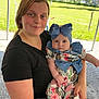 woman, baby, child, outdoor, tent, greenery, grass, person, holding, bow, floral_outfit, blue_bow, headwear, smile, casual_clothing, portrait, daylight, nature, family, candid
