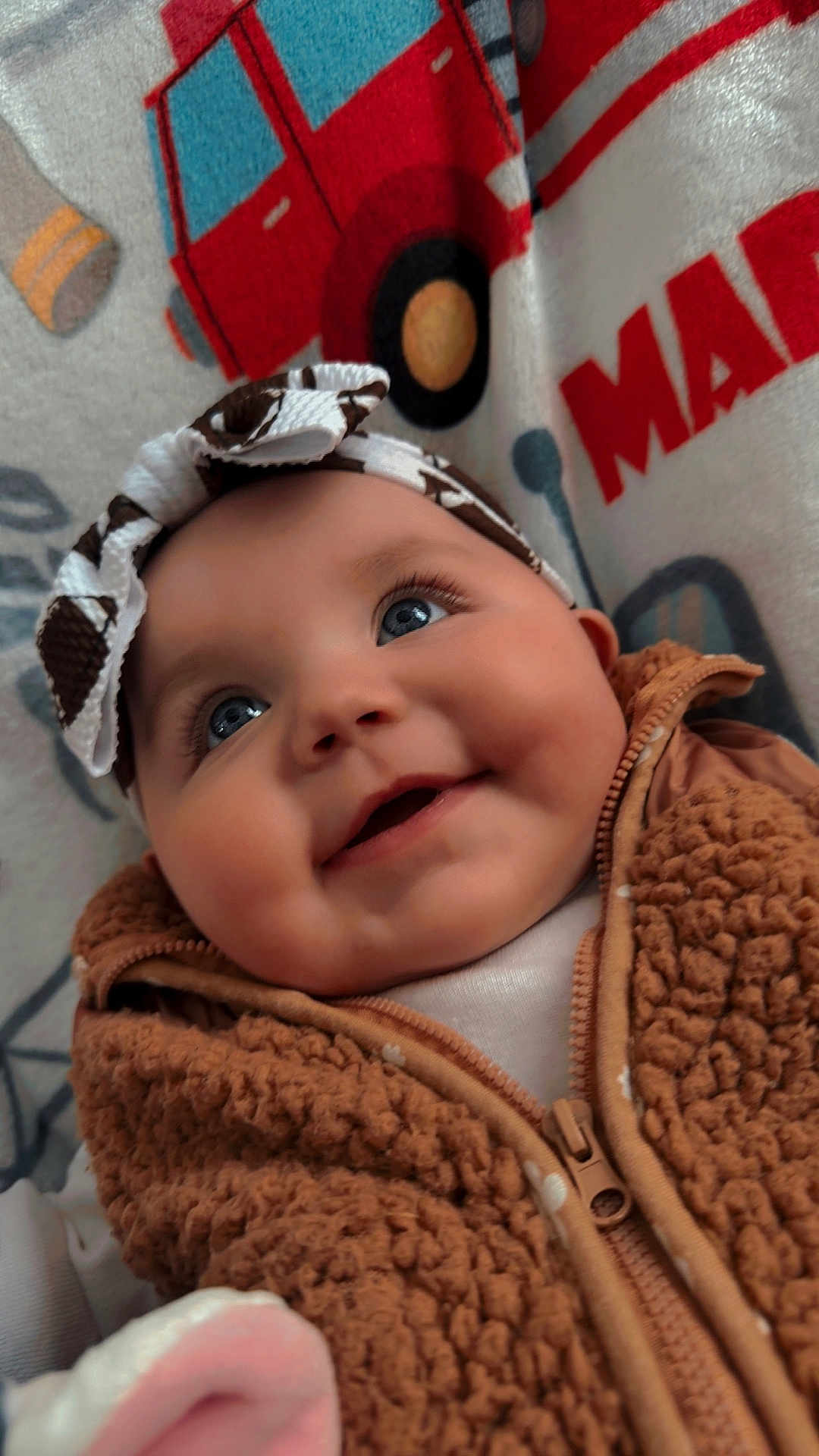 Madeline is registered to the contest to win money with this photo: baby, infant, smiling, blue_eyes, headband, bow, blanket, vehicle_print, jacket, zipper, fleece, close_up, portrait, long_eyelashes, rosy_cheeks, cheeky_smile, soft_texture, indoor, cute, nursery_blanket