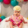 baby, infant, child, portrait, sitting, smiling, wide_eyes, flower_headband, dress, pink_clothing, balloons, birthday, celebration, colorful, indoor, blanket, bed, baby_photography, festive, cute