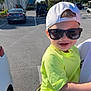 Alaina is registered to the contest to win money with this photo: toddler, child, sunglasses, cap, smile, yellow_shirt, camo_shorts, car, parking_lot, palm_trees, sunny, outdoor, person, happy, casual_clothing, daylight, vehicle, road, shadow, urban