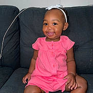 Kyleigha is registered to the contest to win money with this photo: baby, child, clothing, couch, cute, dark_background, face, foot, girl, hand, happy, headband, indoor, infant, person, pink_dress, portrait, sitting, skin, smiling