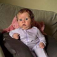 Lyra joined the competition — help win amazing prizes! baby, child, curly_hair, purple_onesie, cushion, couch, pink_blanket, surprised_expression, indoor, face, person, sitting, wide_eyes, infant, young_child, soft_lighting, home_interior, portrait, clothing, hand