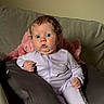 baby, child, curly_hair, purple_onesie, cushion, couch, pink_blanket, surprised_expression, indoor, face, person, sitting, wide_eyes, infant, young_child, soft_lighting, home_interior, portrait, clothing, hand