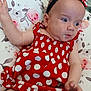 baby, child, clothing, cute, drool, face, floral_pattern, hand, headband, indoor, infant, person, pillow, polka_dot, portrait, red_dress, sitting, skin, smile, young_child