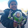 Jeremiah is registered to the contest to win money with this photo: blue_sky, buzz_lightyear, car, child, daylight, face, hand, happy, hat, holding, jacket, outdoor, person, plastic_toy, portrait, sidewalk, smiling, toy, urban, winter_clothing
