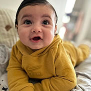 Kaylan a rejoint le concours — aidez-le/la à gagner de superbes lots ! baby, child, hoodie, yellow_clothing, smiling, face, portrait, lying_down, indoors, bed, blanket, cute, happy, person, infant, young_child, cozy, soft_light, closeup, curious