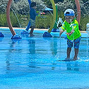 Samuel Davis is registered to the contest to win money with this photo: children, child, splash_pad, water, fountain, outdoor, summer, sunny, playground, rainbow_arches, swimming, wet, smiling, boy, hat, green_shirt, shorts, water_play, park, daylight