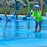 children, child, splash_pad, water, fountain, outdoor, summer, sunny, playground, rainbow_arches, swimming, wet, smiling, boy, hat, green_shirt, shorts, water_play, park, daylight