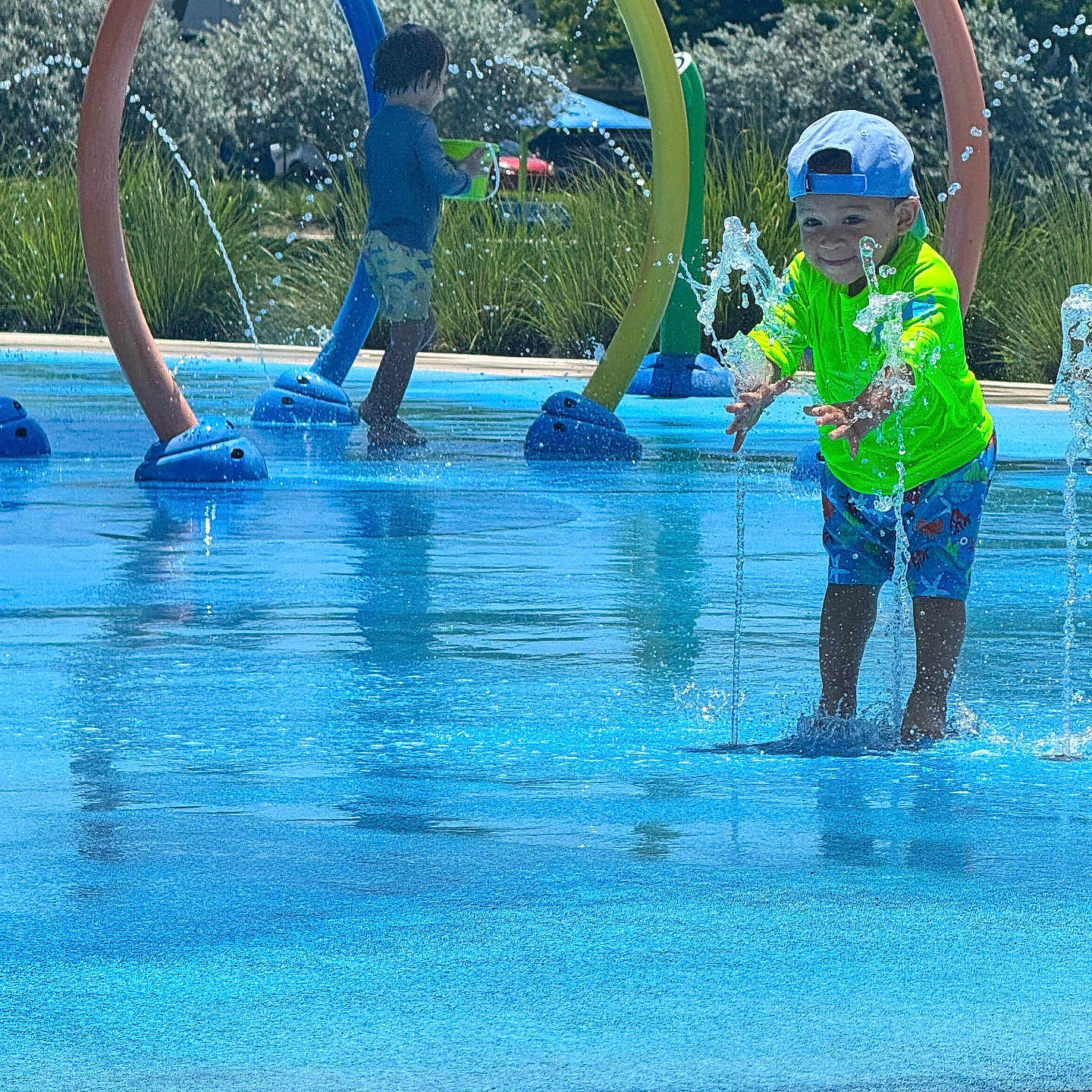 Samuel Davis is registered to the contest to win money with this photo: children, child, splash_pad, water, fountain, outdoor, summer, sunny, playground, rainbow_arches, swimming, wet, smiling, boy, hat, green_shirt, shorts, water_play, park, daylight