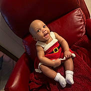 Yalani is registered to the contest to win money with this photo: baby, infant, child, red_chair, knit_dress, white_socks, smiling, happy, seated, indoors, plush, holiday_outfit, cute, portrait, young_child, cozy, red_blanket, skin, face, person