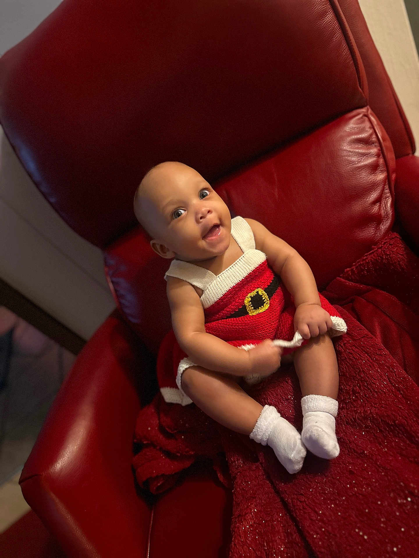 Yalani is registered to the contest to win money with this photo: baby, infant, child, red_chair, knit_dress, white_socks, smiling, happy, seated, indoors, plush, holiday_outfit, cute, portrait, young_child, cozy, red_blanket, skin, face, person