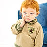child, toddler, red_hair, blue_eyes, sweater, woodland_tree, hand_on_face, green_pants, indoor, portrait, cute, cozy_clothing, expression, curly_hair, seated, soft_lighting, furniture, white_background, casual, young_child