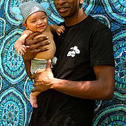 Zaqurian is registered to the contest to win money with this photo: baby, child, man, person, smile, holding, crochet, hat, outdoor, nature, pattern, fabric, blue, green, shirt, black_clothing, happy, portrait, background, casual