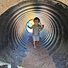 child, toddler, barefoot, tunnel, corrugated_metal, sand, playground, daylight, shadow, clothing, dinosaur_pattern, adventure, outdoor, smiling, person, young_child, play, fun, curved_surface, light