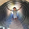 Ares is registered to the contest to win money with this photo: arms_outstretched, barefoot, child, circular_tunnel, corrugated_tunnel, daylight, happy, metal, outdoor, play, playground, sand, shadow, shirt, shorts, smiling, standing, sunlight, toddler, young_child