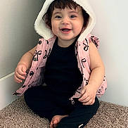 Abby is registered to the contest to win money with this photo: baby, child, smiling, hooded_vest, pink_clothing, black_clothing, carpet, sitting, happy, cute, indoors, toddler, barefoot, face, person, portrait, young_child, casual_clothing, infant, apparel