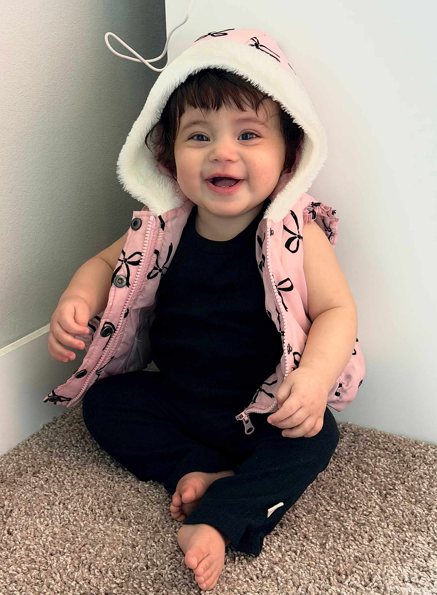 Abby is registered to the contest to win money with this photo: baby, child, smiling, hooded_vest, pink_clothing, black_clothing, carpet, sitting, happy, cute, indoors, toddler, barefoot, face, person, portrait, young_child, casual_clothing, infant, apparel