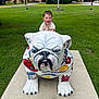 child, toddler, statue, bulldog, grass, park, outdoor, building, tree, sidewalk, happy, smiling, colorful, sculpture, concrete, playful, daytime, person, nature, greenery