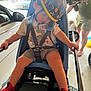 child, helmet, bike_seat, red_shoes, sleeping, straps, shorts, tshirt, adult, car, garage, side_mirror, person, footwear, outdoor, daylight, safety, transportation, relaxation, rest