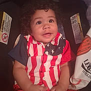 Legend is registered to the contest to win money with this photo: baby, blue, car_seat, child, clothing, curly_hair, cute, happy, indoors, infant, person, portrait, red, seat_belt, shirt, sitting, smiling, stars, stripes, white