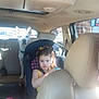 child, toddler, car_seat, dog, white_dog, car_interior, backseat, seatbelt, yellow_clothing, serious_expression, vehicle, window, sunlight, pet, animal, person, transportation, headrest, seat, daylight