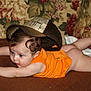 baby, infant, child, blue_eyes, curly_hair, orange_onesie, cap, hat, tummy_time, crawling, couch, floral_wallpaper, blanket, brown_surface, indoor, portrait, curious_expression, bare_legs, plush_fabric, young_child