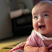 Rylee is registered to the contest to win money with this photo: baby, infant, smiling, tummy_time, drool, bib, pink_clothing, closeup, portrait, cheeks, big_eyes, happy, face, indoor, blanket, blurred_background, soft_lighting, crawling, playmat, young_child