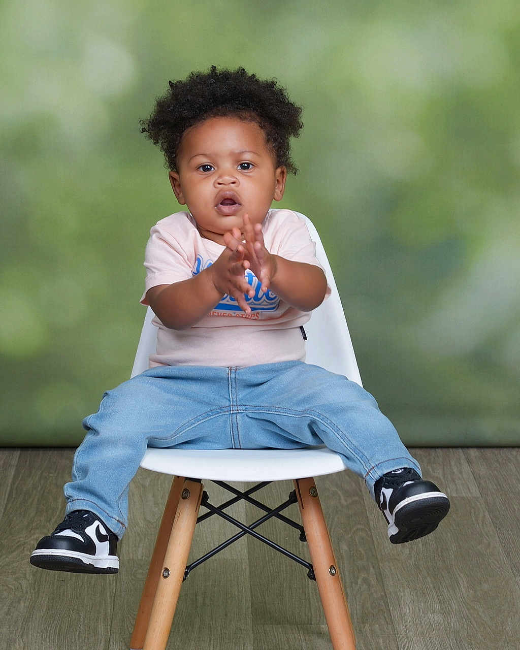 Josiah is registered to the contest to win money with this photo: toddler, child, person, sitting, clapping, chair, white_chair, jeans, tshirt, sneakers, black_and_white_shoes, curly_hair, indoor, wooden_floor, green_background, portrait, cute, young_child, happy, casual_clothing