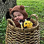 baby, child, costume, bear_costume, basket, plush_toy, bee_toy, outdoor, grass, tree_trunk, nature, cute, smile, face, person, infant, greenery, wicker, sitting, expression
