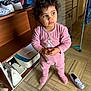 child, toddler, pink_onesie, curly_hair, standing, indoor, shoe_rack, slipper, shoes, wooden_shelf, floor_tiles, portrait, hands, expression, laundry_rack, cabinet, walking_shoes, home_interior, cute, looking_away