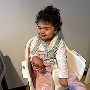 Arya participe au concours pour gagner de l'argent avec cette photo : child, toddler, curly_hair, highchair, harness, straps, smile, snack, hand, coat, long_sleeve, pants, indoor, wall, seat, wooden_leg, portrait, happy, baby_chair, cute