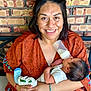 Lucian is registered to the contest to win money with this photo: woman, baby, feeding, bottle, smile, brick_wall, holding, socks, clothing, arm, happy, person, infant, portrait, indoor, casual, hair, furniture, necklace, baby_clothes