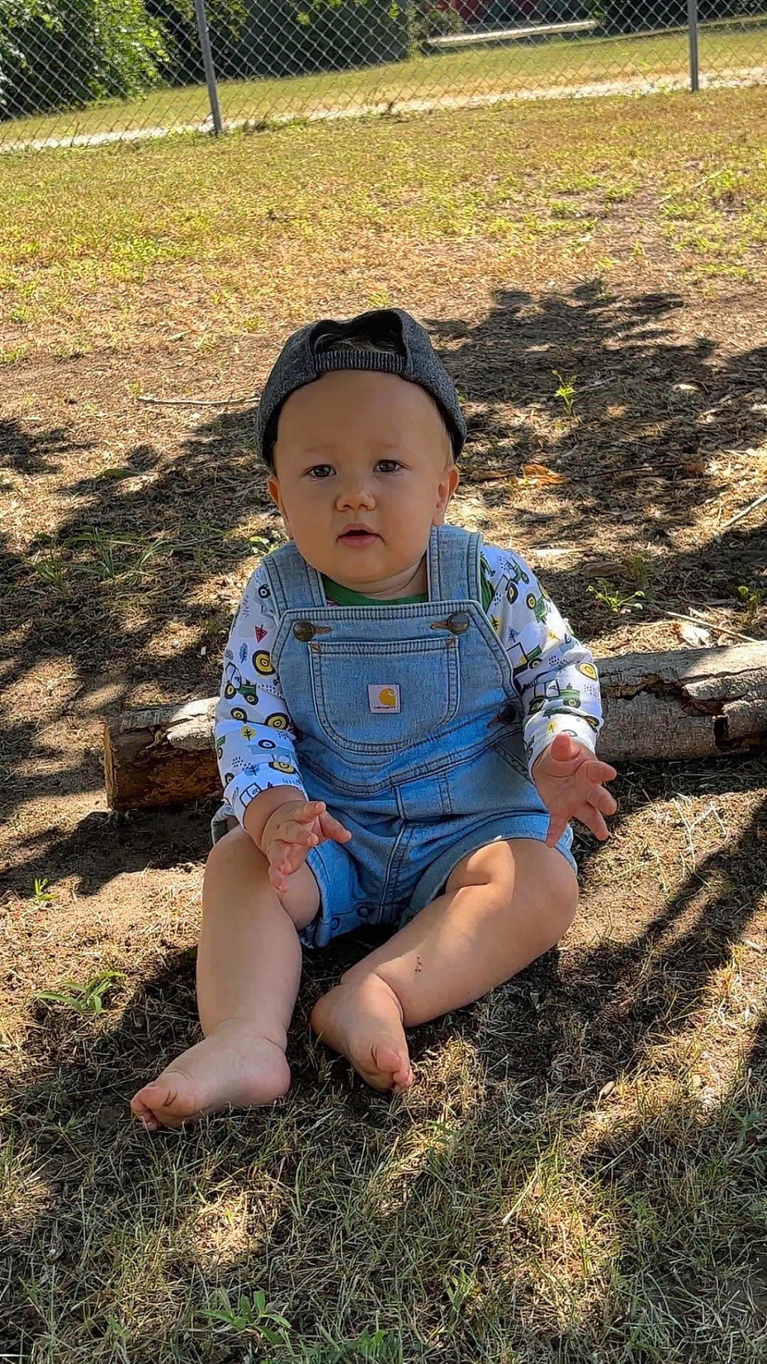 Lucas is registered to the contest to win money with this photo: child, toddler, baby, overalls, cap, barefoot, grass, sunlight, shadow, outdoor, log, fence, curious, sitting, nature, clothing, face, person, summer, playful